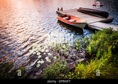 Sunset riflettendo in acqua di lago con canotto legato al dock in legno a spiaggia rocciosa. In Ontario, Canada. Foto Stock