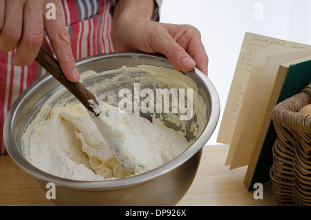 Mani torta di agitazione mescolare con il cucchiaio di legno accanto al libro di ricette. Foto Stock