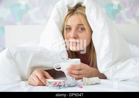 Ritratto di giovane donna con tazza da caffè e medicinali affetti da febbre mentre coperto di panno sul letto Foto Stock