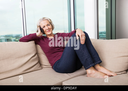 Lunghezza intera di senior donna rispondendo alle smart phone sul divano di casa Foto Stock