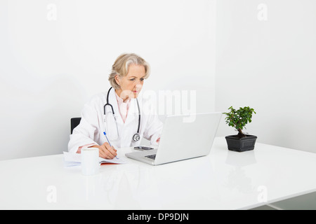Senior medico donna scrivere note mentre guarda il computer portatile in clinica Foto Stock
