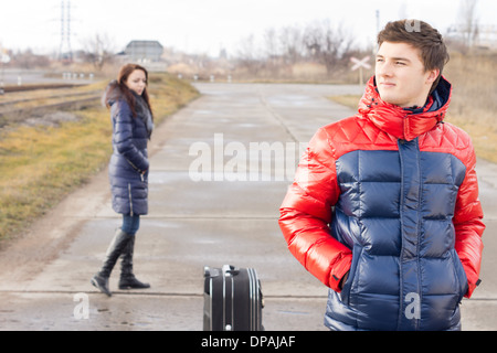 Bel giovane in attesa in strada con una valigia pazientemente in piedi con le mani in tasca mentre un yong donna cammina da dietro di lui. Foto Stock