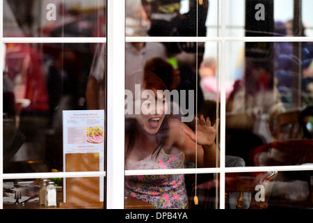 Piuttosto giovane donna sat in un ristorante sventolare a un amico visto attraverso la finestra Foto Stock