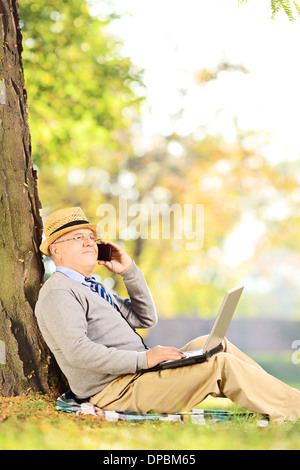 Senior uomo nel parco a parlare su un telefono cellulare e di lavoro sul computer portatile Foto Stock