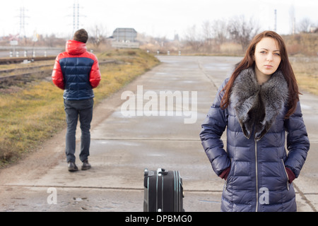 Giovane donna in attesa con la sua valigia per un sollevamento in mezzo alla strada mentre un giovane uomo cammina passato nella direzione opposta. Foto Stock