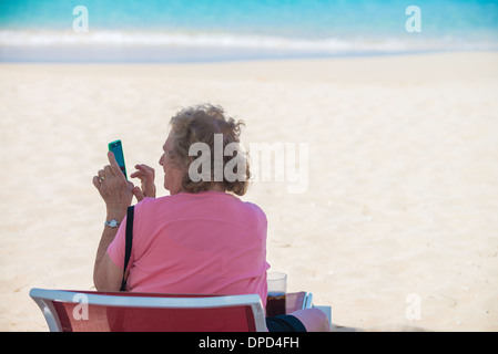 Un'anziana donna caucasica si siede in una sdraio sulla spiaggia sulla spiaggia di Sandcastle, affacciato sul Mar dei Caraibi e gioca con il suo smart phone. Foto Stock