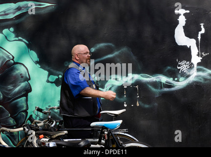 Uomo in divisa dalla parete graffiti dietro una bicicletta spazio parcheggio Brick Lane area, East London E1 REGNO UNITO Foto Stock