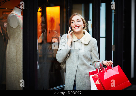 Giovane donna camminare per strada portando shopping bag e rendendo chiamata telefonica Foto Stock