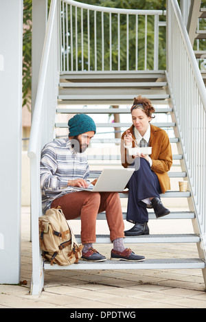 Hipster giovane utilizzando il computer e mangiare il pranzo seduti in scale al campus universitario Foto Stock