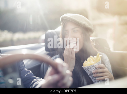 Coppia giovane nel convertibile, donna mangiare patatine Foto Stock