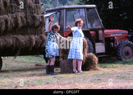 Due donne gallesi più anziane che sventolano le mani lavorando in un'azienda agricola caricando balle di fieno su un trattore e un rimorchio durante la produzione del fieno nel 1984 1980 nel Carmarthenshire Galles Regno Unito Gran Bretagna KATHY DEWITT Foto Stock