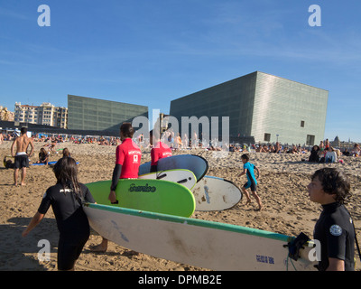 Surfers e lucertole da mare sulla spiaggia Zurriola con centro congressi Kursaal e auditorium in background, San Sebastian, Spagna Foto Stock