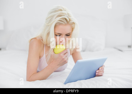 Bionda informale di mangiare un Apple durante l'utilizzo di tablet PC A LETTO Foto Stock