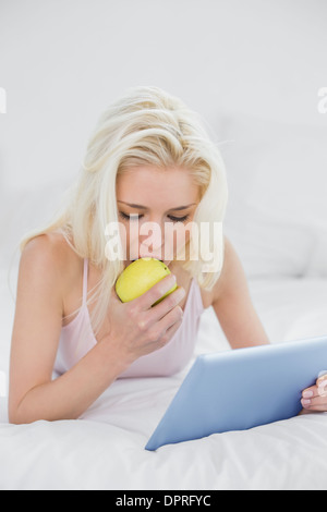 Bionda informale di mangiare un Apple durante l'utilizzo di tablet PC A LETTO Foto Stock
