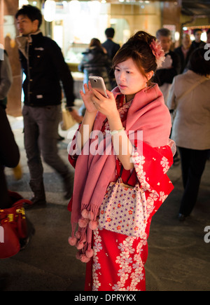 Giovane ragazza giapponese indossa un fiorito inverno rosso kimono in una fresca serata autunnale, mentre utilizzando uno smartphone. Foto Stock