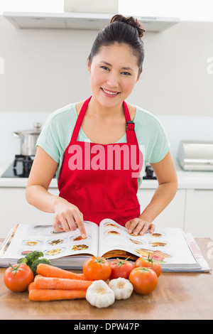 Donna sorridente con il libro di ricette e verdure in cucina Foto Stock