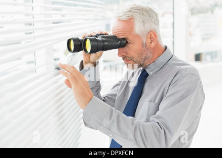 Imprenditore peeking con il binocolo attraverso persiane in office Foto Stock