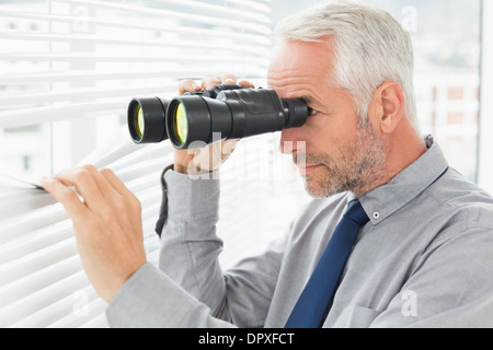 Imprenditore peeking con il binocolo attraverso persiane Foto Stock