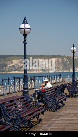 Uomo seduto su un banco di lavoro quotidiano di lettura e mangiare banana a Swanage pier nel Maggio Foto Stock