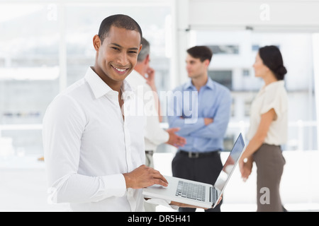 Giovane uomo d affari con computer portatile con il team dietro di lui sorridente in telecamera Foto Stock