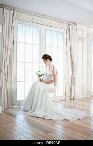 Una donna seduta su una sedia con abito da sposa e bouquet di fiori Foto Stock