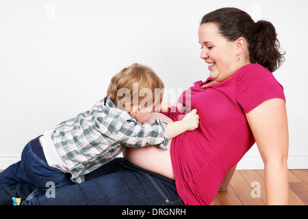 Grazioso piccolo ragazzo biondo cercando baby il fratello o la sorella di sua madre in stato di gravidanza la pancia Foto Stock