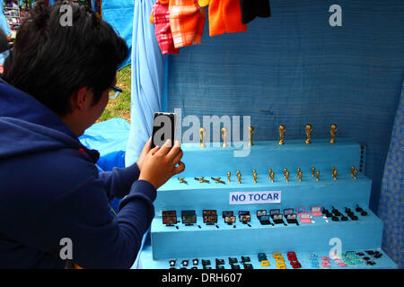 LA PAZ, Bolivia, 26 gennaio 2014. Una persona che prende una foto del mondo in miniatura tazze in vendita presso il festival Alasitas con il suo telefono cellulare dotato di fotocamera digitale. Al festival di persone acquista miniature di ciò che si desidera ottenere nel corso dell'anno e li benedisse in modo che i loro desideri si avverano. Miniature popolari disponibili includono vetture, case, denaro, grado di certificati, computer portatili e televisori (come in questa foto); gli oggetti relativi alle attuali eventi di attualità sono anche fatti come la Coppa del mondo per il Brasile 2014. Credito: James Brunker / Alamy Live News Foto Stock