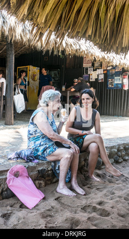 Vecchia donna a mangiare il gelato & spettegolare con la giovane donna seduta relativa sul cordolo di terrazza al famoso ristorante sulla spiaggia Foto Stock