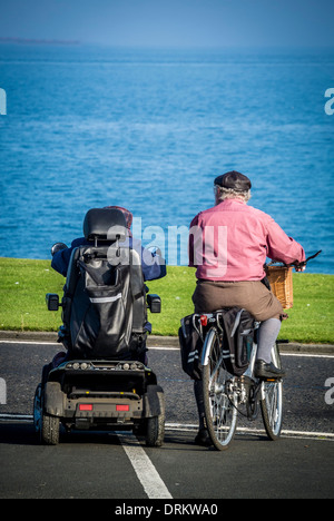 Coppia di anziani su una bicicletta e scooter di mobilità che guarda al mare sulla passeggiata a Whitley Bay. Foto Stock