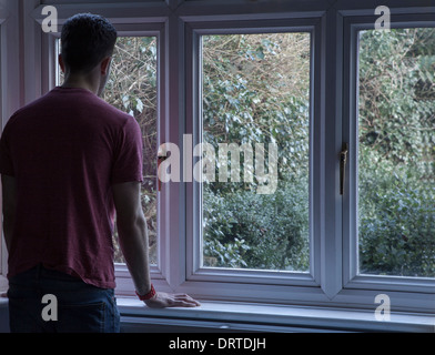 Giovane uomo all'interno guardando fuori attraverso una finestra, sulla spalla vista. Foto Stock