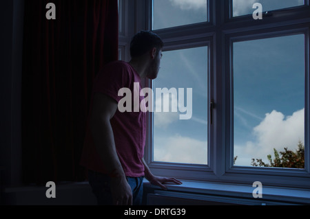 Giovane uomo in una stanza buia guardando fuori attraverso una finestra. Foto Stock