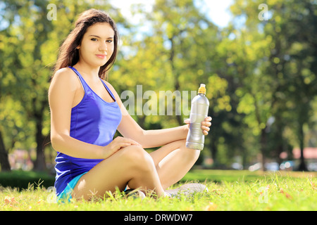 Giovane atleta donna seduta su un'erba dopo exericise e tenendo una bottiglia di acqua in un parco Foto Stock