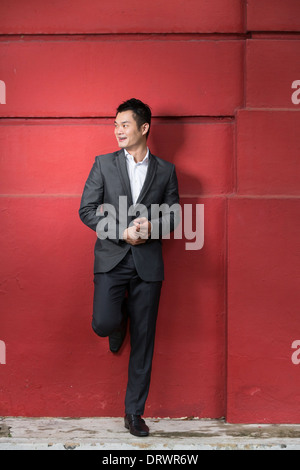 Bel giovane azienda cinese uomo appoggiato contro una parete rossa. Felice Asian business uomo sorridente e guardando lontano. Foto Stock