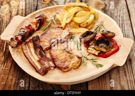 Vista dettagliata del maiale fritto di carne con gli spicchi di patate e verdure alla griglia su un piatto in legno e decorate con rosmarino Foto Stock