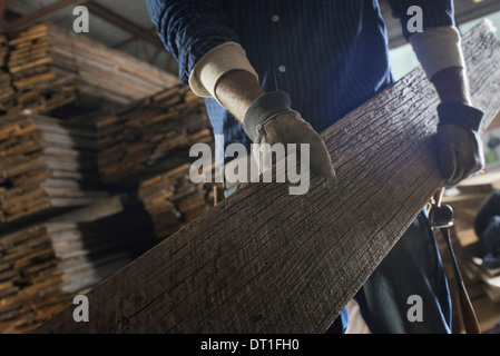 Un cumulo di materiale riciclato rigenerato listoni di legno di legno in un cantiere in legno di un uomo che porta un grande asse di legno Foto Stock