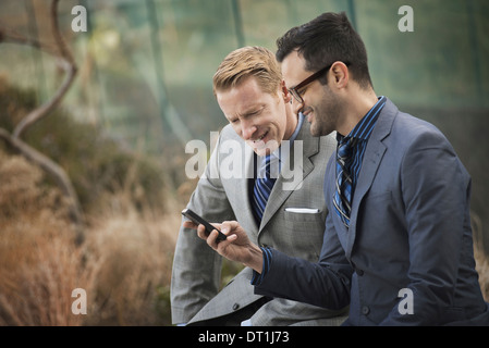 Due uomini in business formale vestiti in piedi a fianco a fianco alla ricerca di un telefono cellulare schermo o telefono cellulare Foto Stock