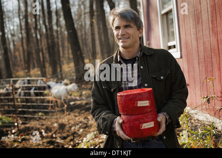 Un uomo in piedi in un cortile di fattoria azienda due blocchi di formaggio di capra Foto Stock