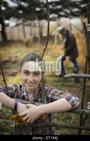 Una giovane ragazza appoggiato su di una recinzione su di una azienda agricola biologica di un uomo di scavare il suolo in background Foto Stock