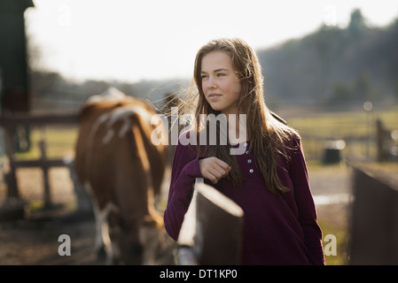 Una ragazza appoggiata su un paddock recinzione e un cavallo al pascolo in background su di una azienda agricola biologica Foto Stock