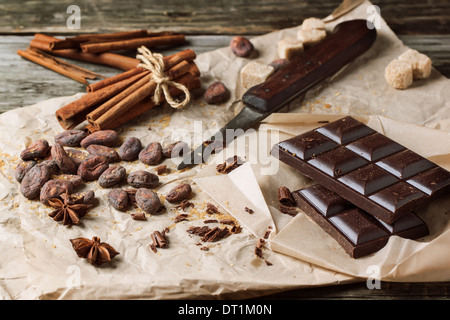 Il cioccolato fondente con il vecchio coltello, le fave di cacao, cannella e anice su carta sgualcita su sfondo di legno Foto Stock