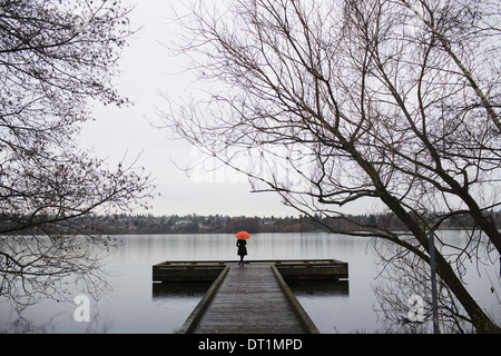 Una donna in piedi alla fine di un dock con un ombrello arancione su un nuvoloso giorno grigio in Seattle Foto Stock