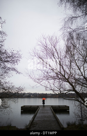 Una donna in piedi alla fine di un dock con un ombrello arancione su un nuvoloso giorno grigio in Seattle Foto Stock