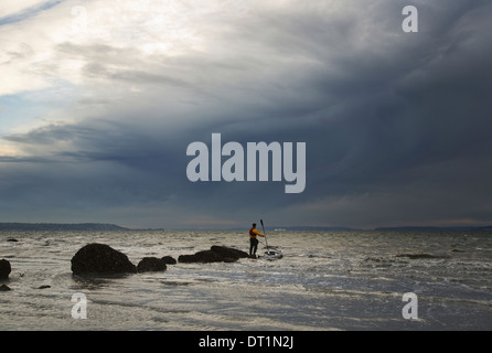 Un uomo si erge su una roccia nel mezzo del Puget Sound con il suo mare kayak floating accanto a lui Foto Stock