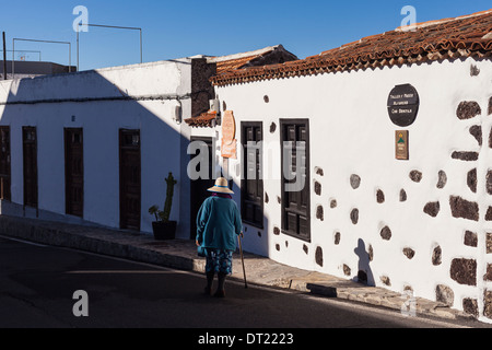 Old Lady con bastone indossando cappello di paglia e blue coat camminando sulla strada di Arguayo, al di fuori del museo della ceramica, Tenerife, Foto Stock