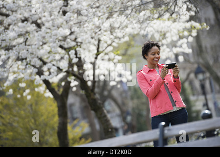 All'aperto nella città in primavera tempo New York City Park fiori bianchi su alberi di una donna che mantiene il suo telefono cellulare e sorridente Foto Stock