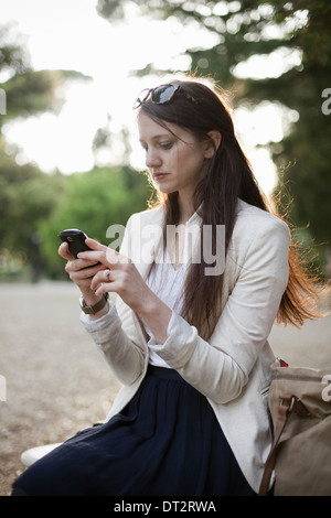Una donna che indossa una giacca di crema seduti in un parco della città guardando il suo telefono cellulare Foto Stock