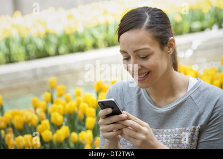 Uno stile di vita urbano una donna nel parco in un letto di tulipani gialli utilizzando il suo telefono cellulare Foto Stock