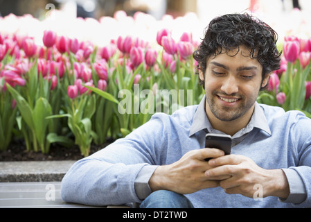 Uno stile di vita urbano un uomo nel parco utilizzando il suo telefono cellulare un letto di rosa fioritura tulipani in background Foto Stock