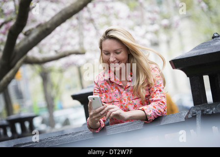 Vista su cityA giovane donna con capelli lunghi biondi seduti in un parco della città guardando il suo smart phone Foto Stock