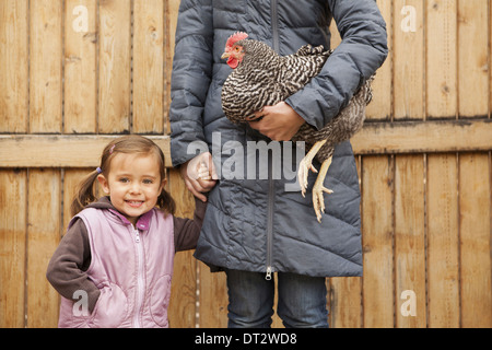 Donna che mantiene un bianco e nero il pollo con un rosso coxcomb sotto un braccio di una giovane ragazza accanto a lei mentre tiene il suo altro lato Foto Stock